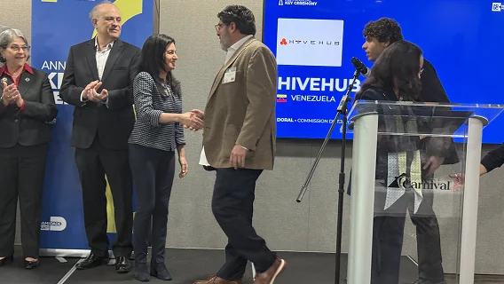 Man in a tan blazer shaking hands with a woman on stage beside a screen showing HiveHub, as attendees applaud