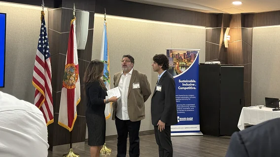 Woman handing papers to a man in a tan blazer beside US, Florida and county flags at a Miami-Dade Beacon Council event